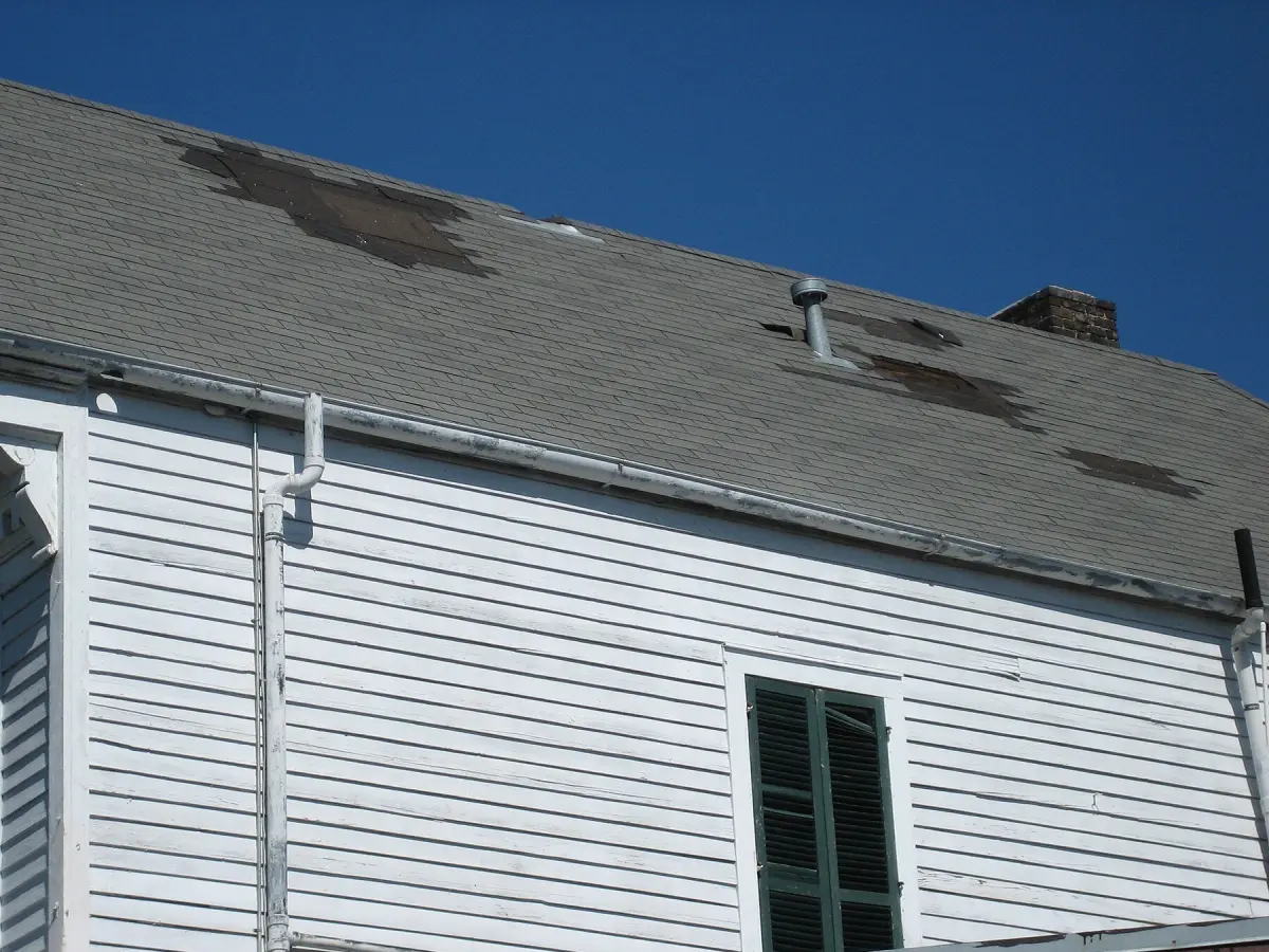 Grey shingle roof on a white clapboard house with patches of shingles torn away by wind, exposing the felt beneath